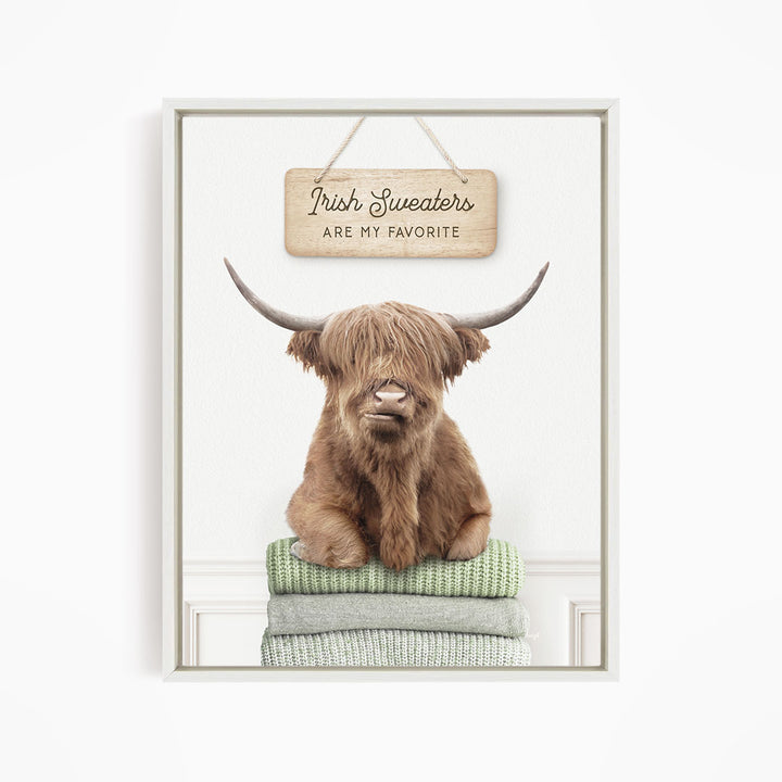A brown Highland cow with large horns is sitting on a stack of green and gray blankets, with a wooden sign above that says "Irish Sweaters are my favorite".