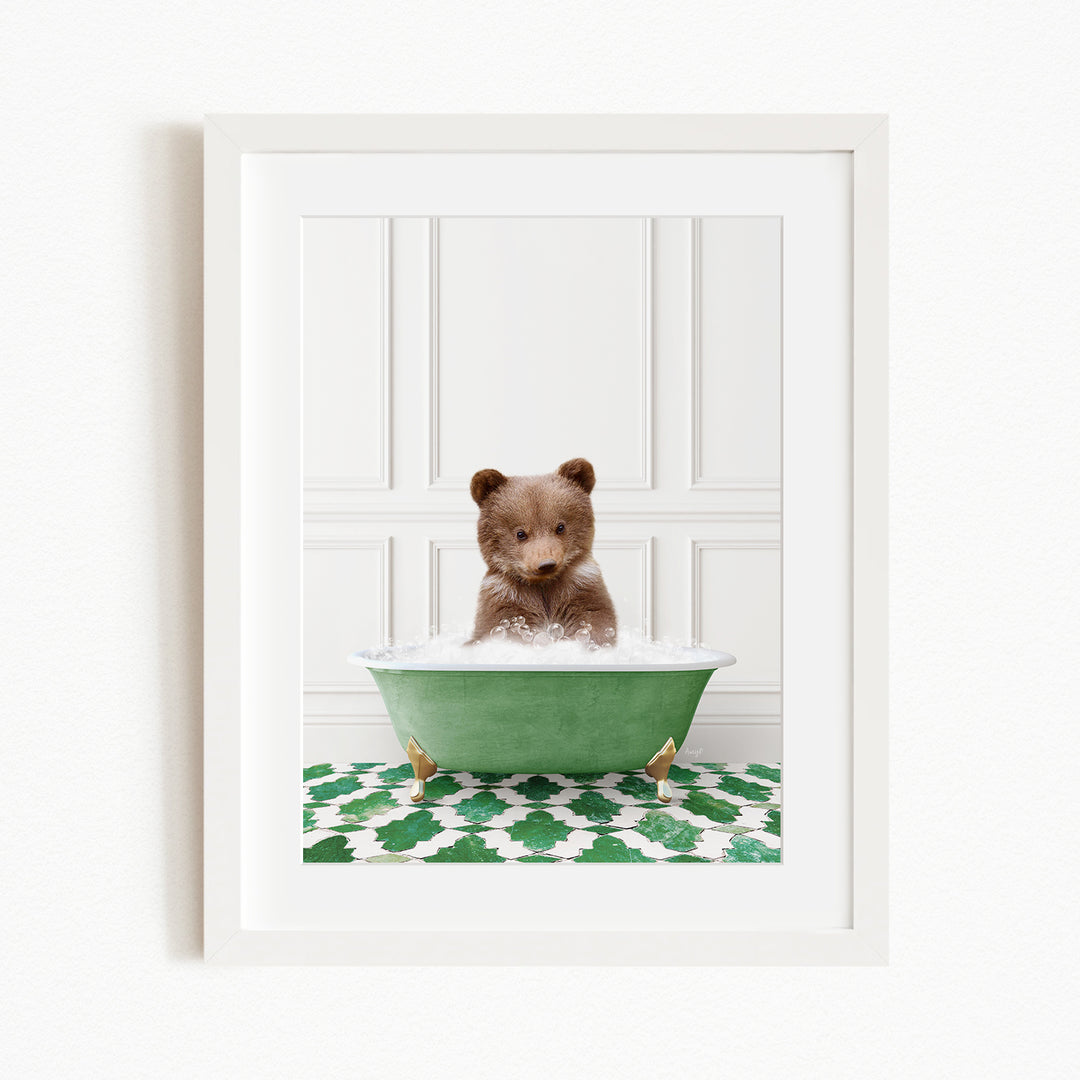 A small brown bear cub sitting in a green bathtub filled with bubbles, surrounded by a green and white patterned rug.