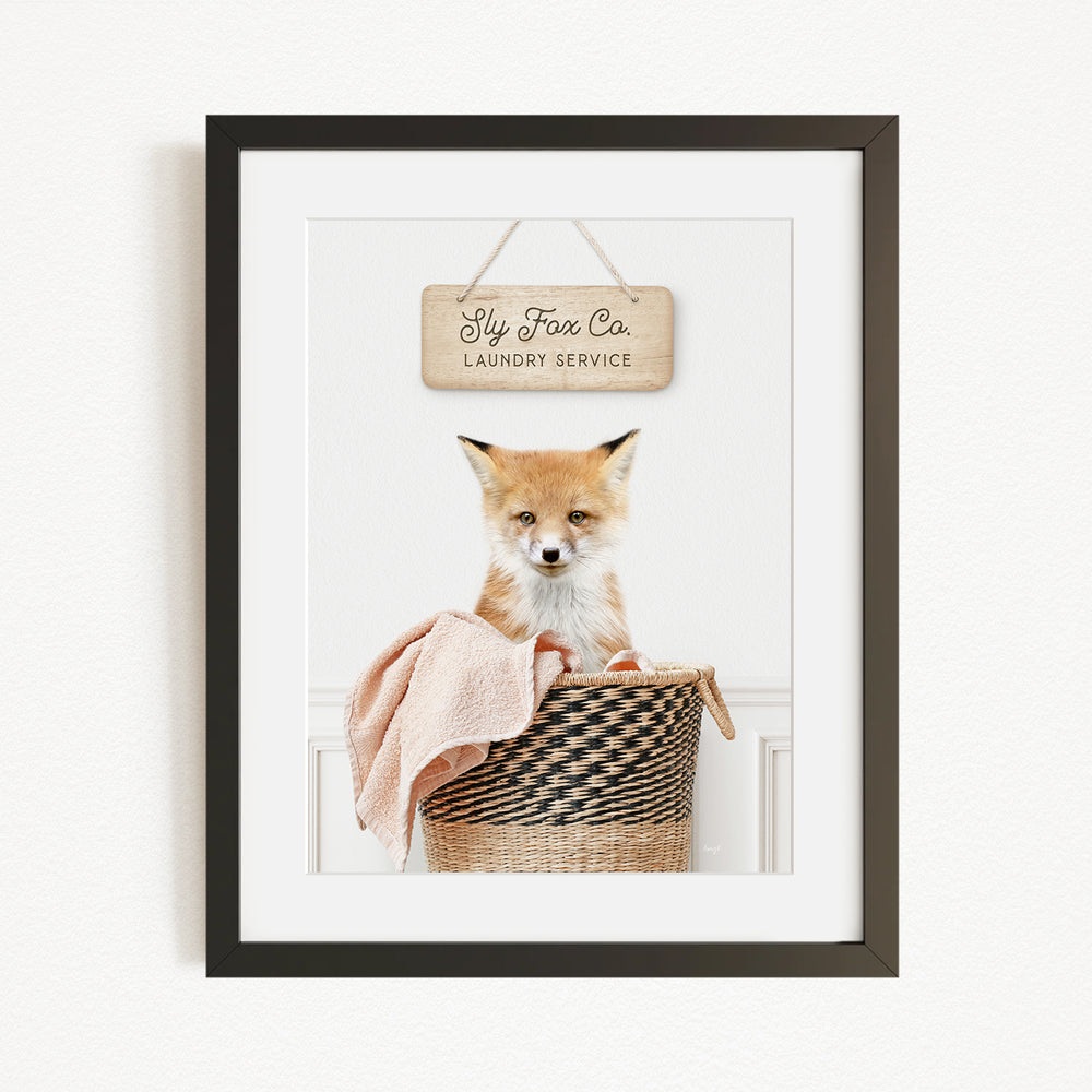 A framed photograph of a young fox sitting in a woven laundry basket, with a sign above that reads "Fly Fox Co. Laundry Service".