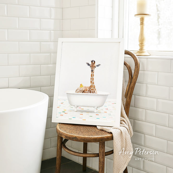A framed image of a giraffe sitting in a white bathtub, with a towel draped over the back of a wooden chair in a bathroom setting.