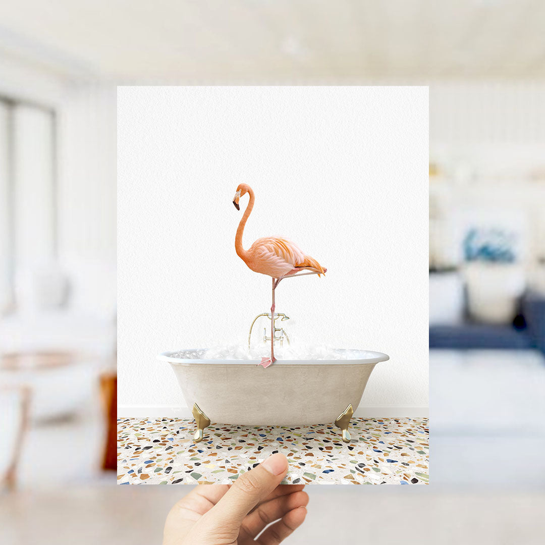 A hand holds up a framed image of a pink flamingo standing on a white bathtub filled with water, creating a whimsical and playful scene.