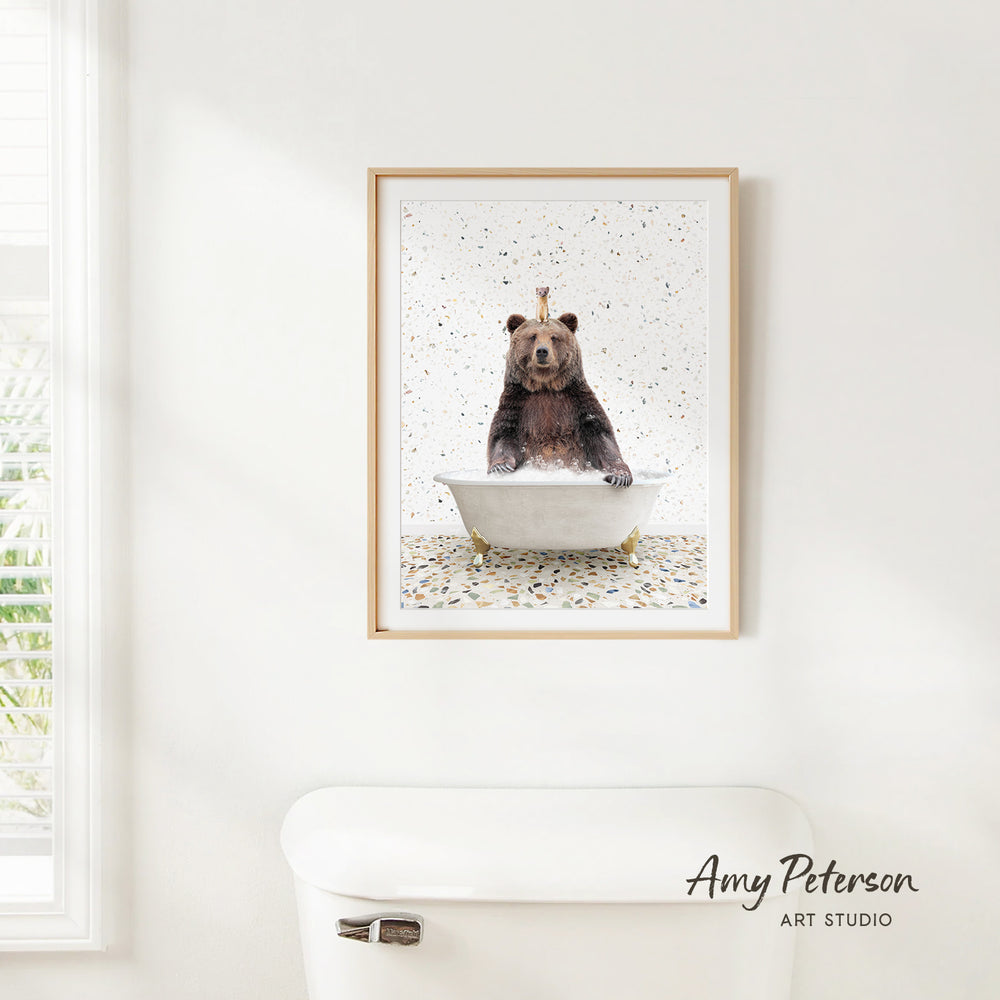 A framed image of a bear sitting in a bathtub, surrounded by confetti, is hanging on a white wall.