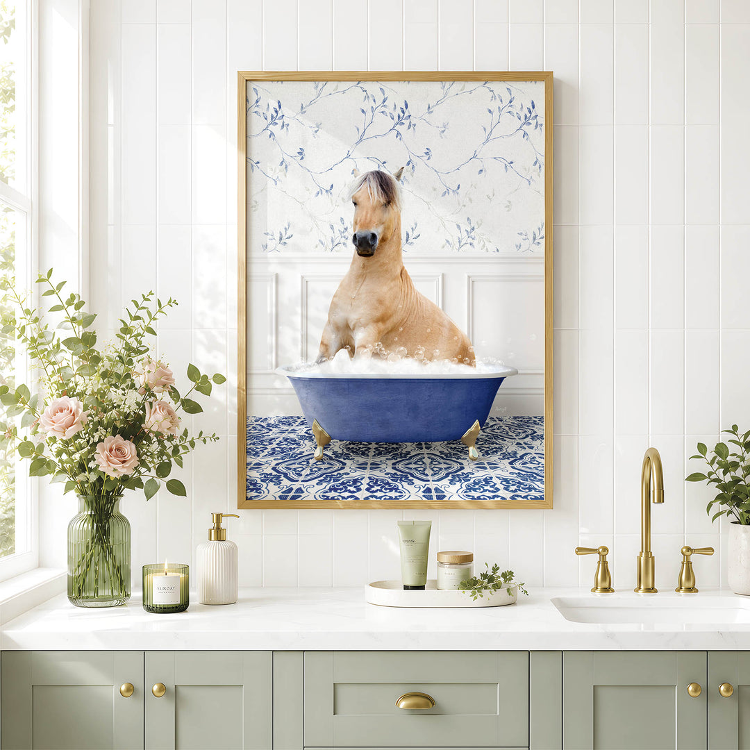 A framed image of a horse sitting in a blue bathtub, surrounded by flowers and a decorative vase, is displayed on a white tiled wall above a bathroom counter with various toiletries and a plant.