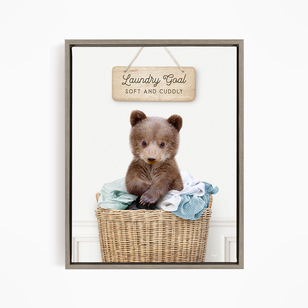 A cute brown bear sitting in a wicker laundry basket, with a wooden sign above that says "Laundry Goal Soft and Cuddly".