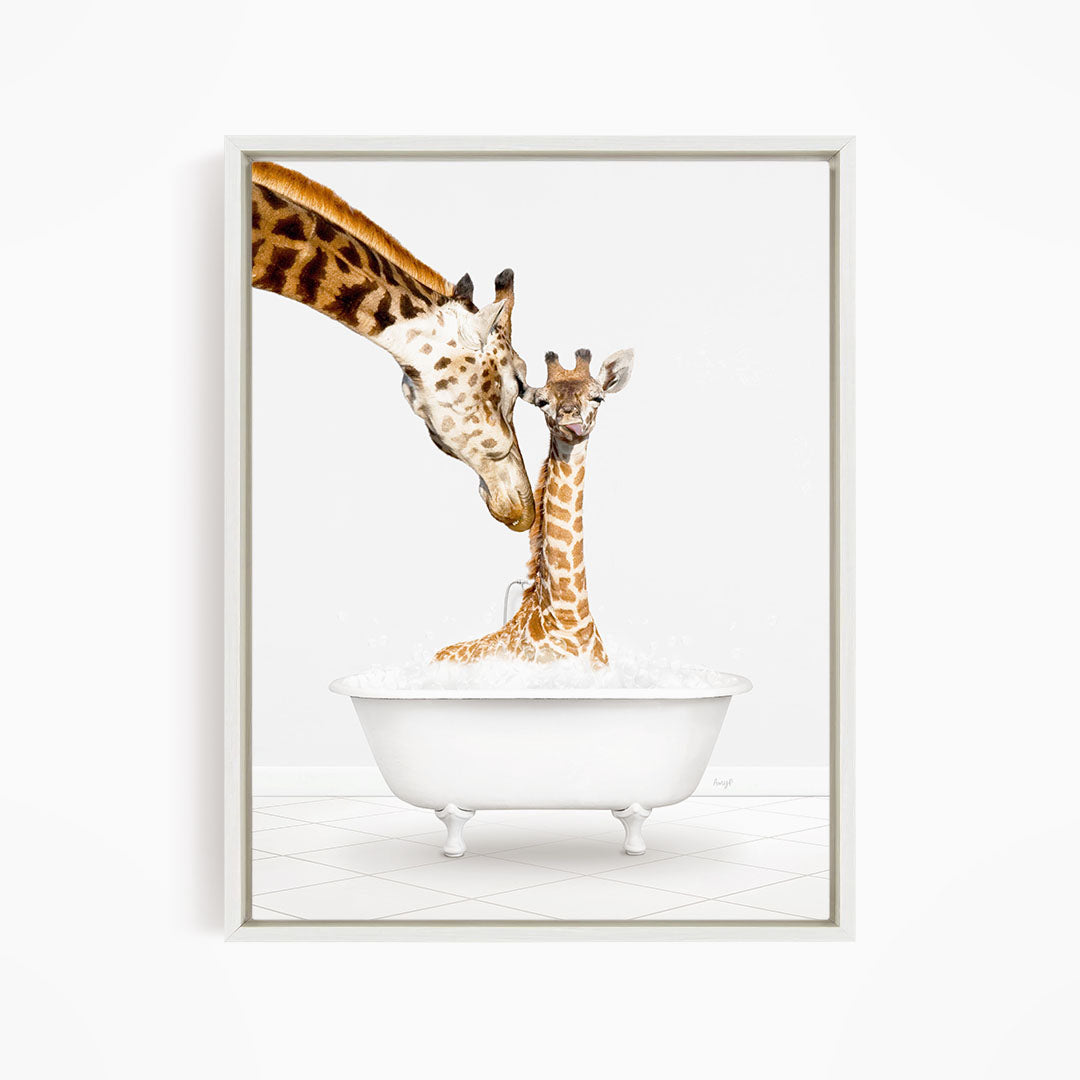 A mother giraffe and her baby giraffe are sitting together in a white bathtub, with the mother gently licking her baby.