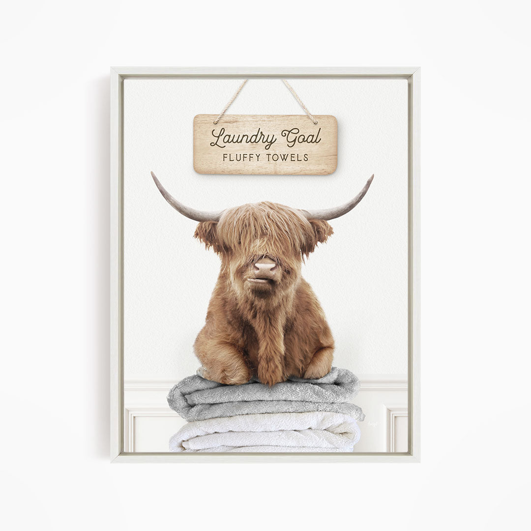 A brown cow with long horns is sitting on a stack of fluffy towels, with a sign above that says "Laundry Goal Fluffy Towels".