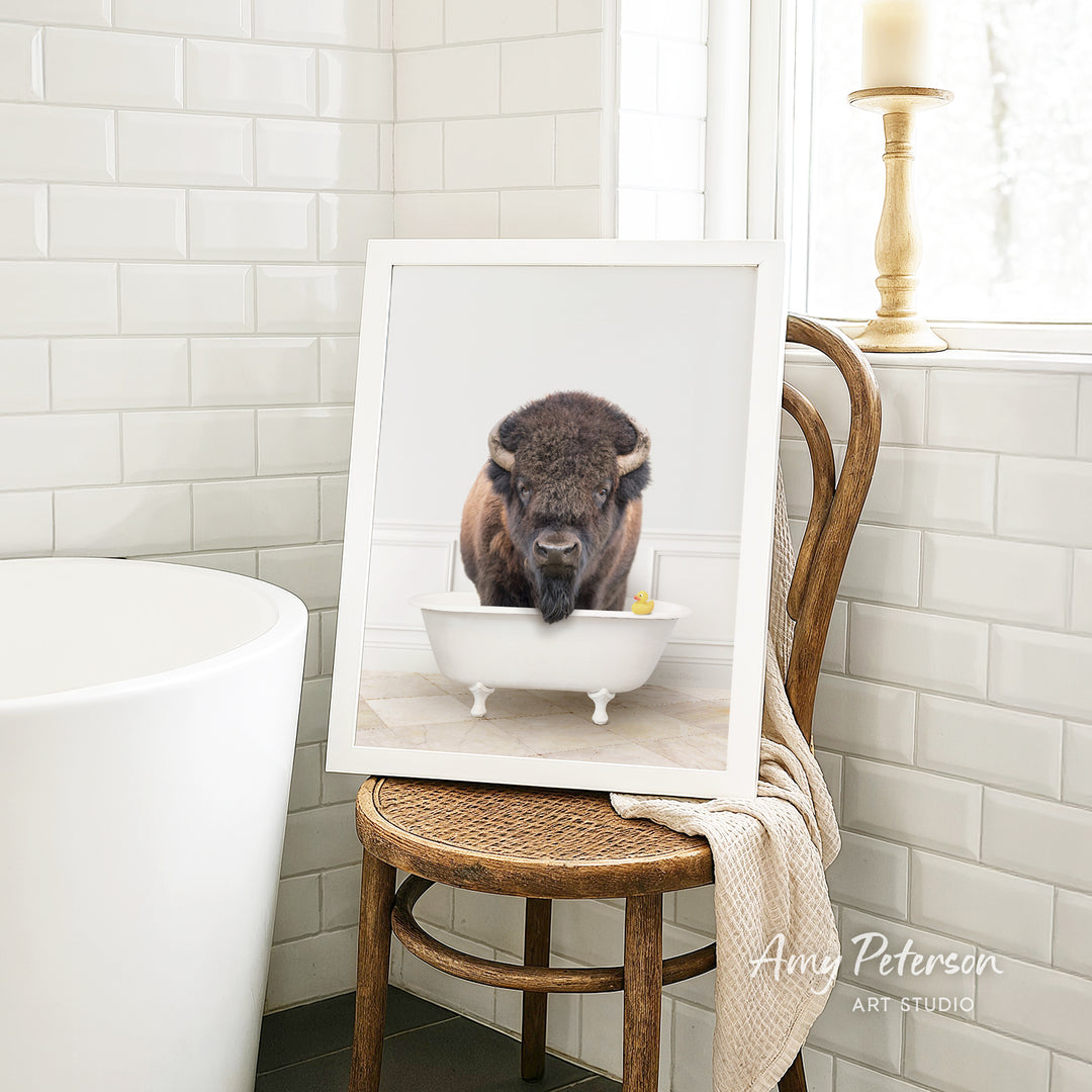 A framed image of a buffalo standing in a white bathtub, with a towel draped over the back of a wooden chair in a bathroom setting.