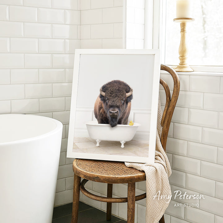 A framed image of a buffalo standing in a white bathtub, with a towel draped over the back of a wooden chair in a bathroom setting.