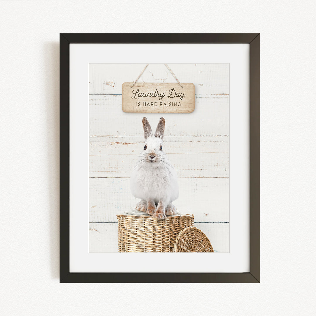 A framed image of a white rabbit sitting on a wicker basket, with a wooden sign above it that says "Laundry Day is here raising".