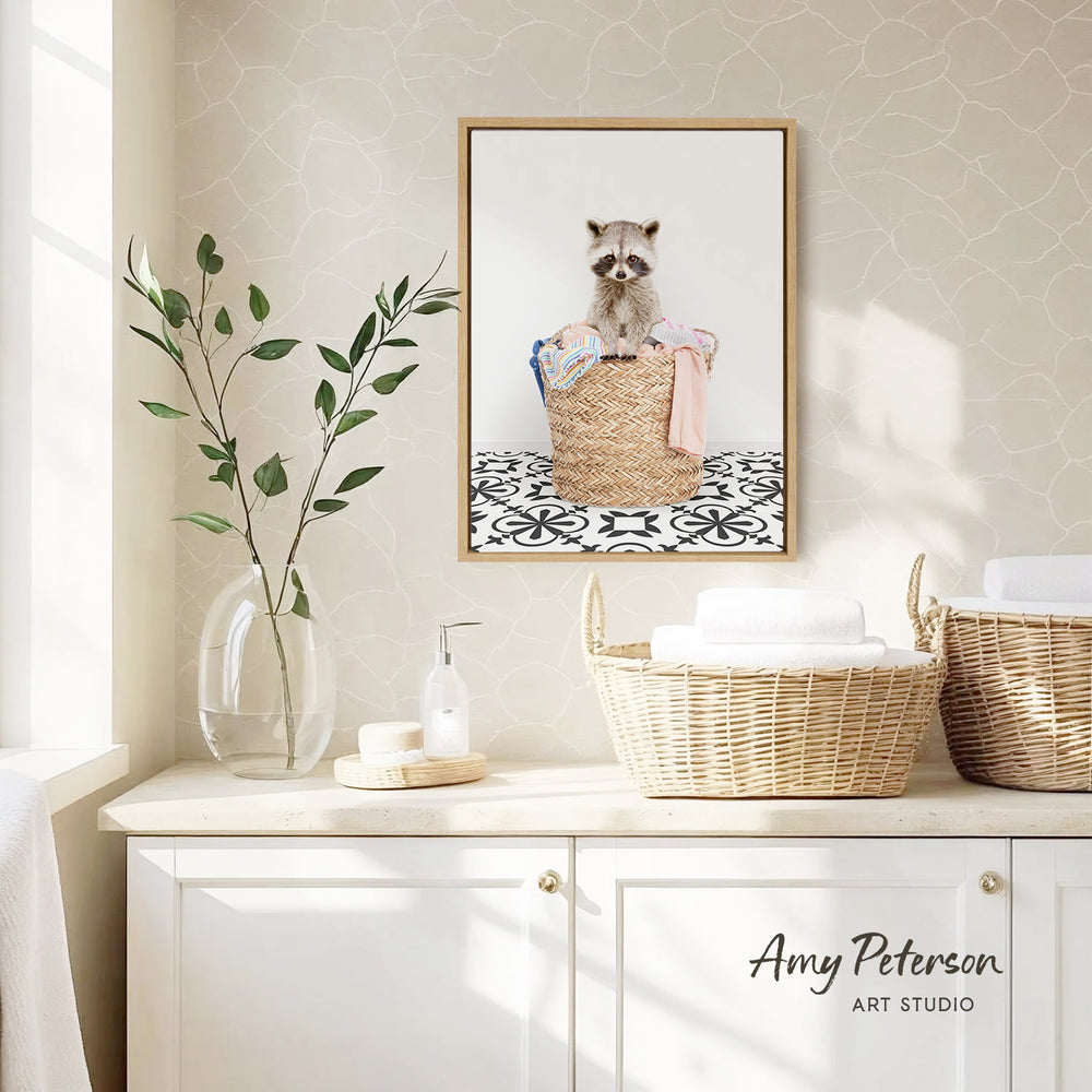 a small raccoon sitting in a woven basket, which is placed on a white countertop. The countertop also has a vase with a plant and a soap dispenser on it. Above the countertop, there is a framed picture of a raccoon in a basket. The wall behind the countertop has a patterned wallpaper.