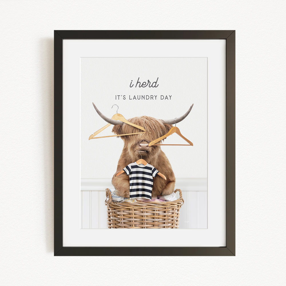A framed image depicting a Highland cow sitting in a wicker basket, with a pair of clothes hangers hanging over its head. The text above the cow reads "It's laundry day".