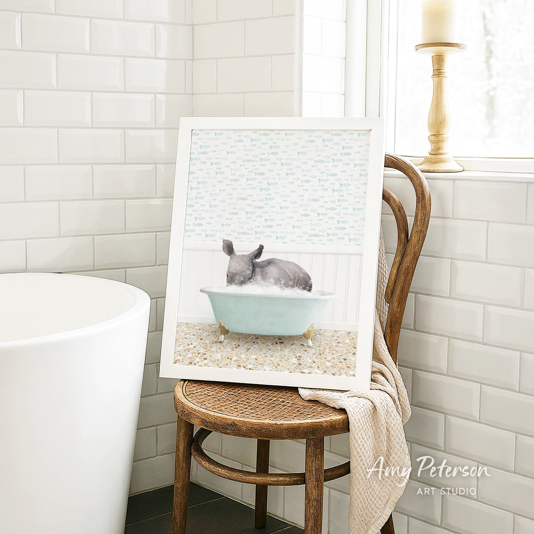 A framed image of a gray rabbit sitting in a blue bathtub, surrounded by a patterned wallpaper, is displayed on a wooden chair in a bathroom setting.
