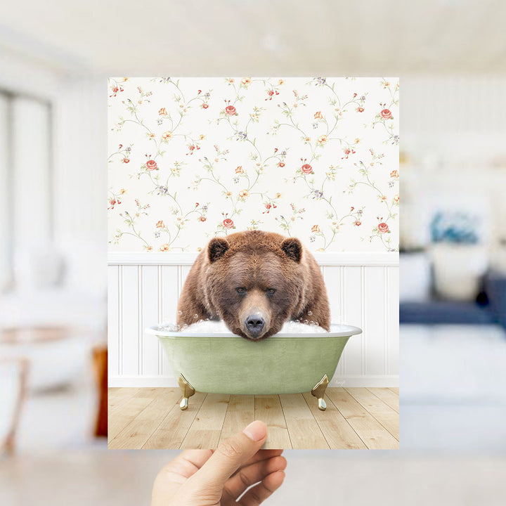 A hand holds up a framed image of a brown bear sitting in a green bathtub, with a floral wallpaper in the background.