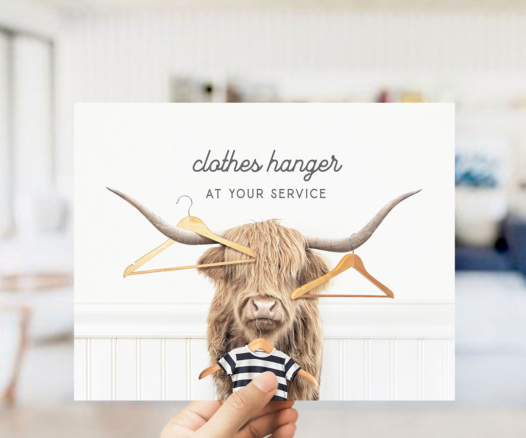 A hand holding a card with a picture of a horned animal, specifically a yak, wearing a striped shirt and holding a hanger. The text on the card reads "clothes hanger at your service".