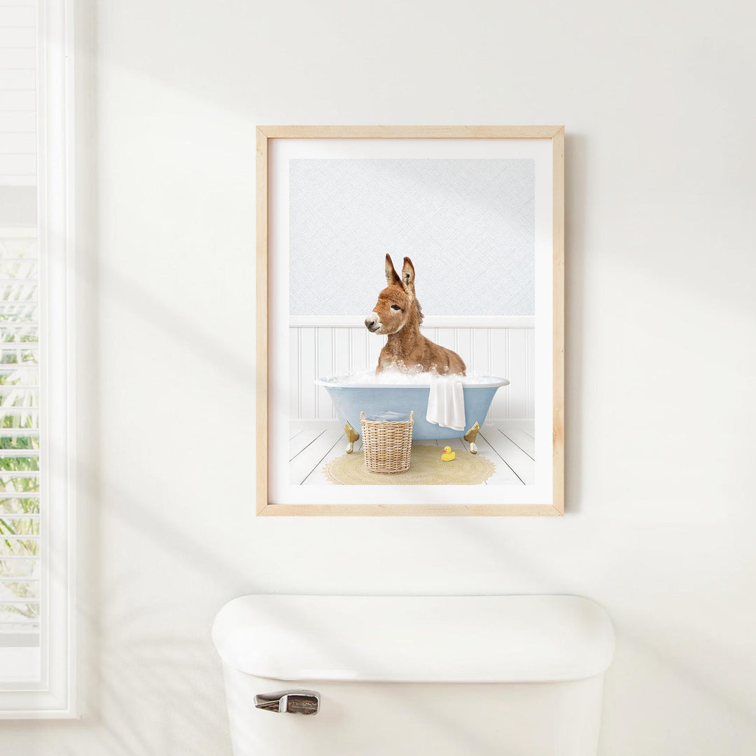 A framed image of a donkey sitting in a bathtub, surrounded by a basket of bath toys and a towel, is displayed on a white wall above a white toilet.
