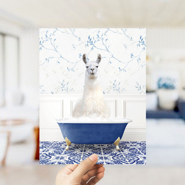 A white llama sitting in a blue bathtub filled with bubbles, with a blue and white floral wallpaper in the background.