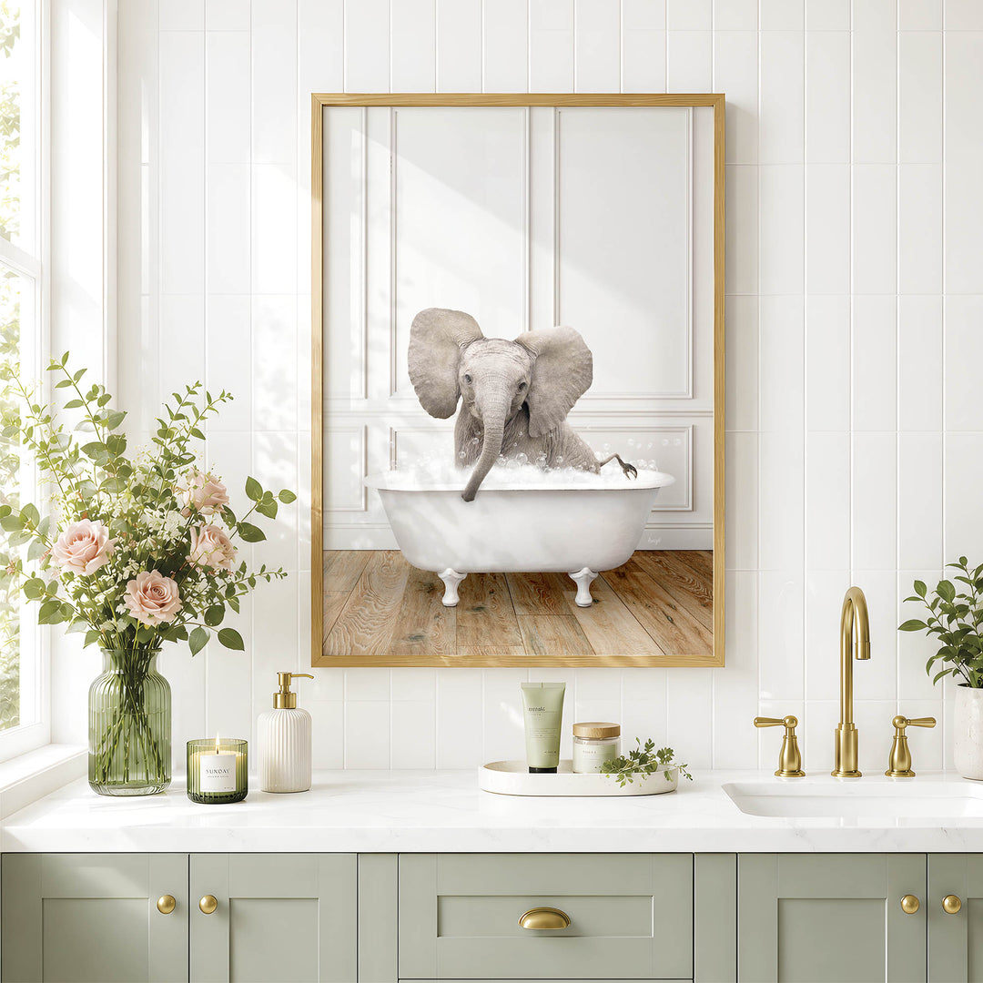 A framed image of an elephant sitting in a bathtub, surrounded by flowers and candles, is displayed on a white tiled wall above a bathroom vanity with a sink and gold faucets.