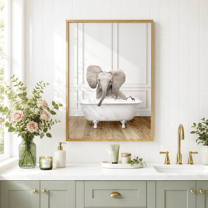 A framed image of an elephant sitting in a bathtub, surrounded by flowers and candles, is displayed on a white tiled wall above a bathroom vanity with a sink and gold faucets.