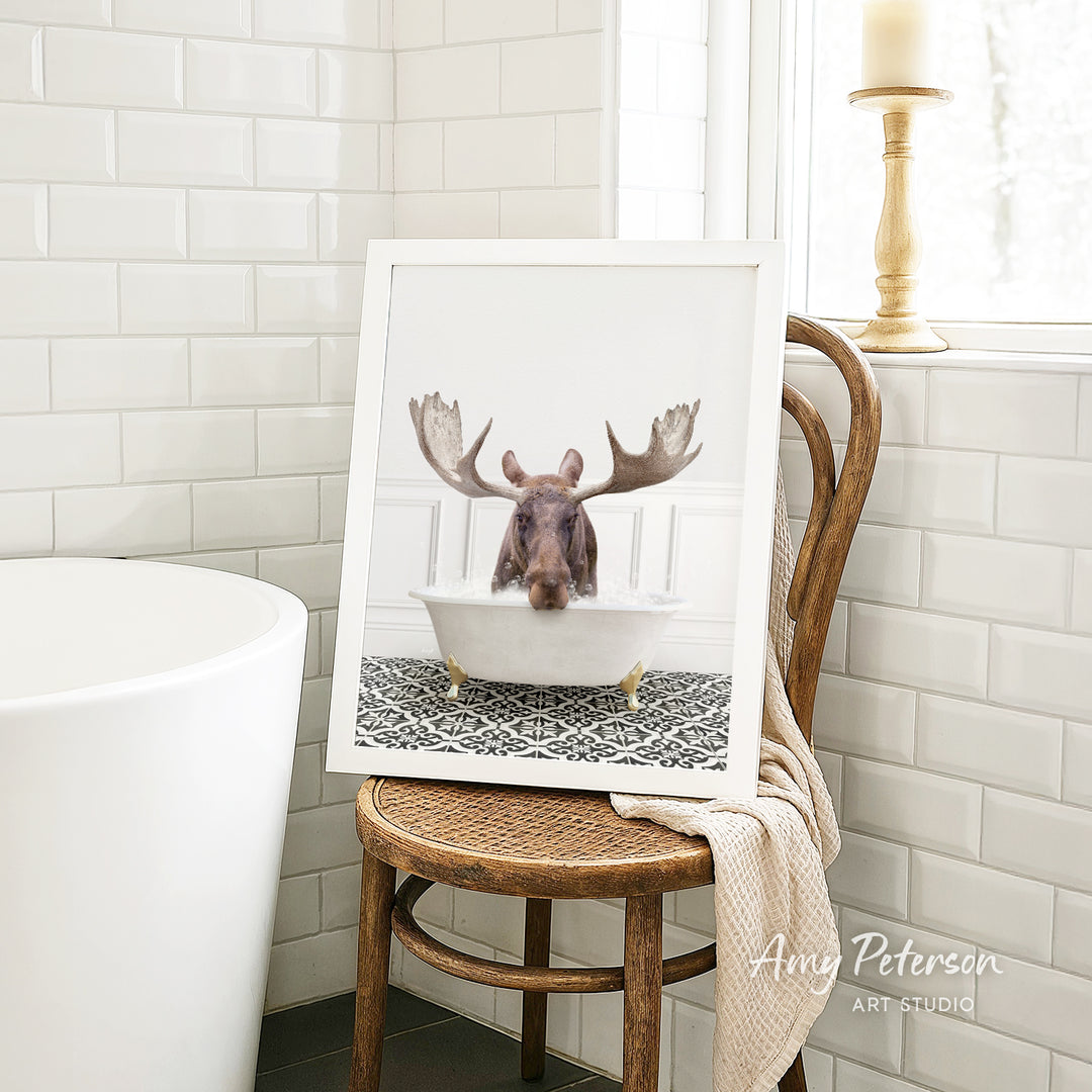 A framed image of a moose sitting in a bathtub is displayed on a wooden chair in a bathroom setting.