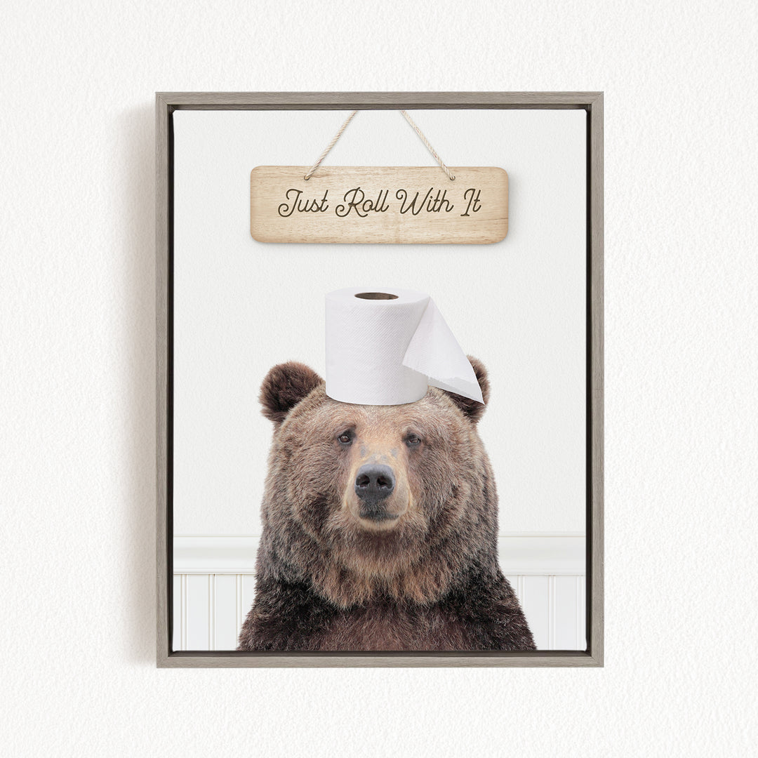 A framed image of a brown bear wearing a toilet paper roll on its head, with a wooden sign above it that says "Just Roll With It".