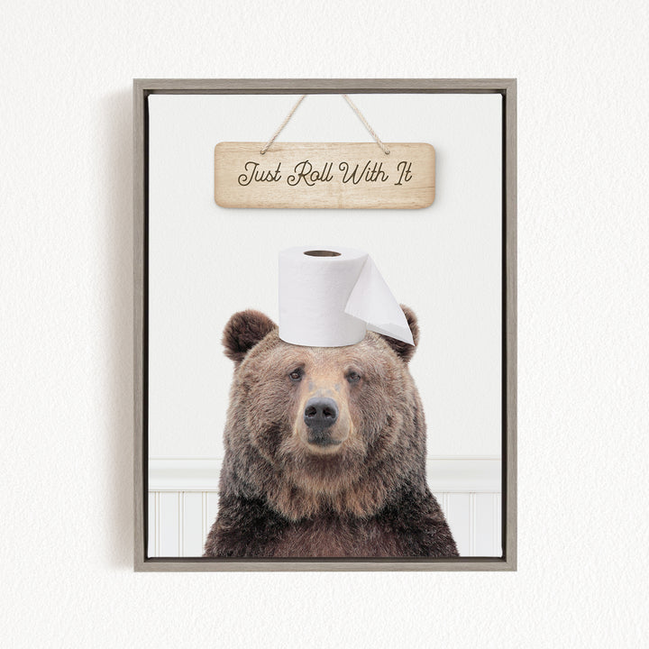 A framed image of a brown bear wearing a toilet paper roll on its head, with a wooden sign above it that says "Just Roll With It".