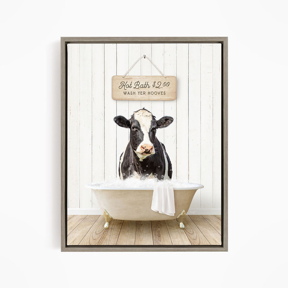 A black and white cow is sitting in a white bathtub, with a wooden sign above it that reads "Hot Bath $2.00 WASH YOUR HOoves".