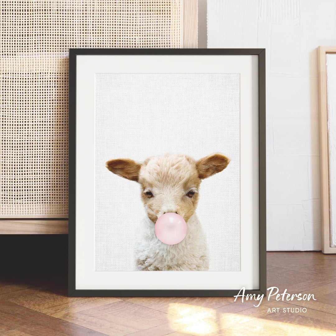 A framed image of a lamb blowing a bubble gum bubble, with a wooden floor and a woven basket in the background.
