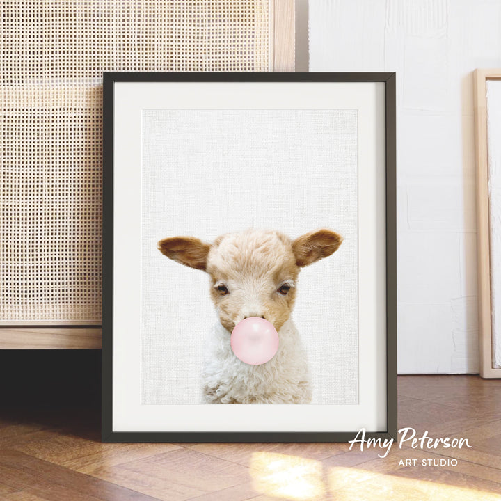A framed image of a lamb blowing a bubble gum bubble, with a wooden floor and a woven basket in the background.