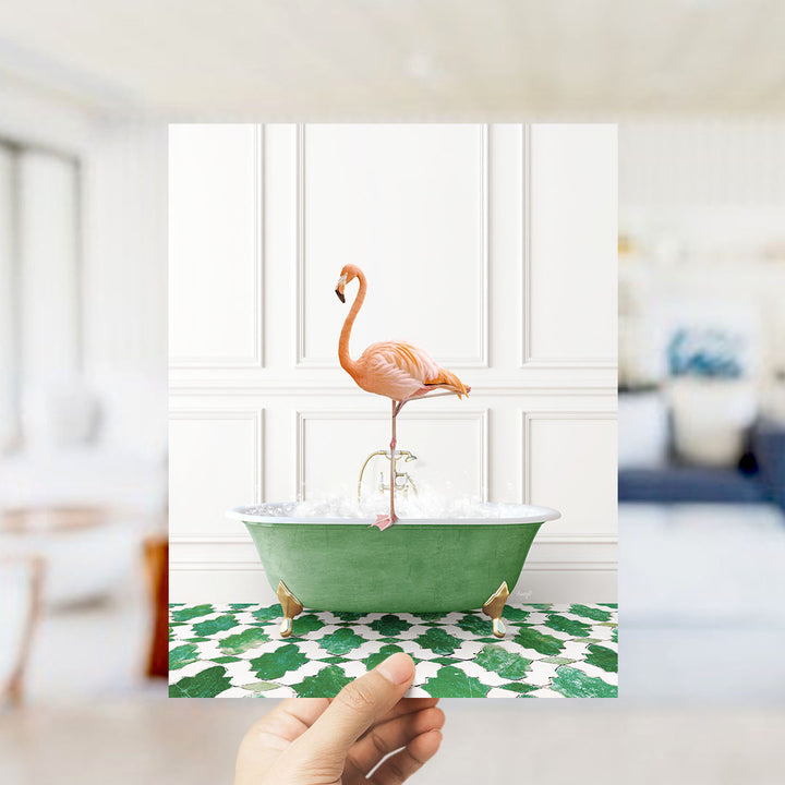 A hand holds up a framed image of a pink flamingo standing on a green bathtub filled with water, set against a white wall with a green and white patterned floor.