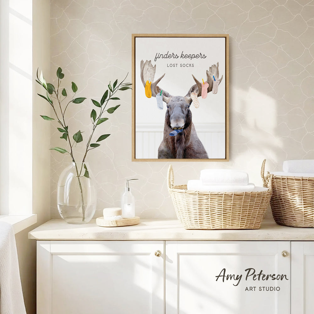 a framed poster of a moose with a humorous caption, "Finders keepers: Lost socks", hanging on a wall above a white cabinet with baskets.