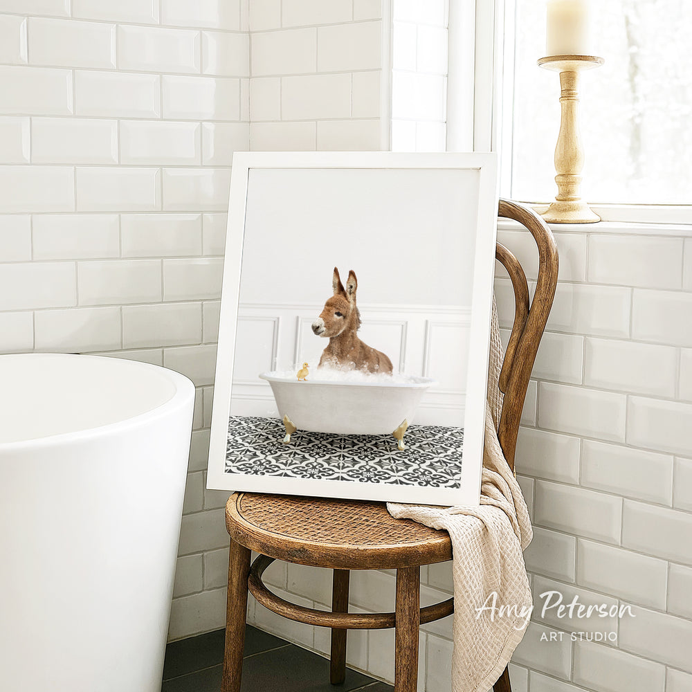 A framed image of a small dog sitting in a bathtub, surrounded by a patterned tile design, is displayed on a wooden chair in a bathroom setting.