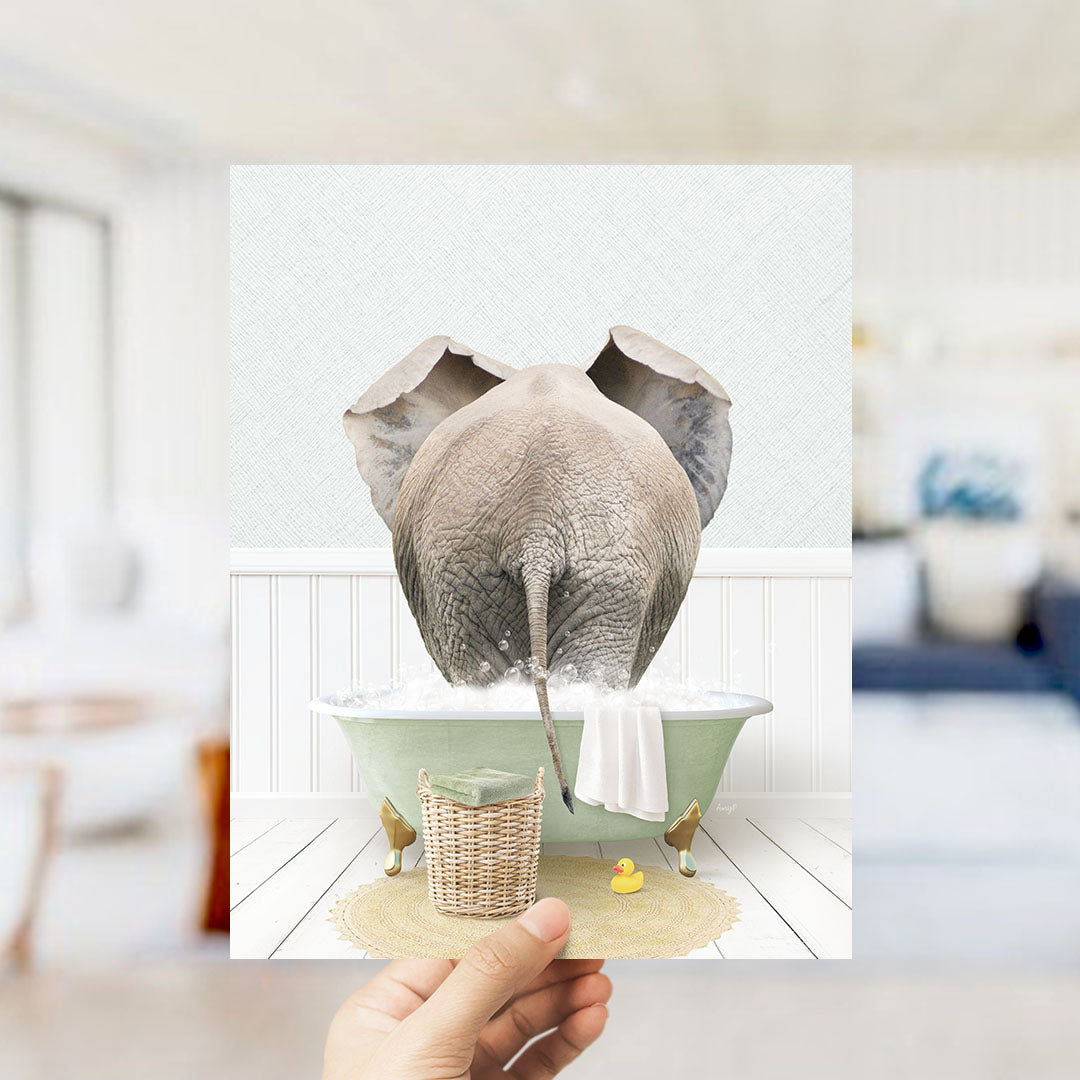 A hand holds up a framed image depicting an elephant standing in a bathtub filled with water, surrounded by a yellow rubber duck and a white towel.