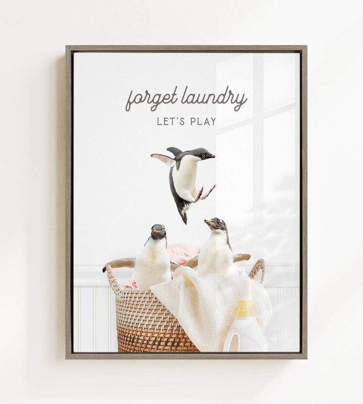 A framed image depicting three penguins in a basket, with the text "forget laundry, let's play" written above them.