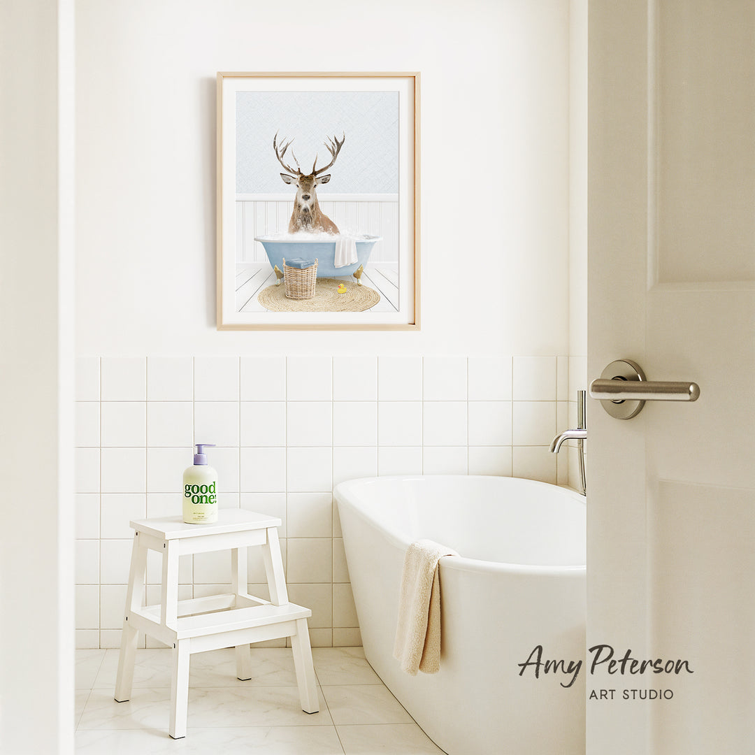 A framed image of a deer sitting in a bathtub is displayed on the wall above a white bathtub in a bathroom.