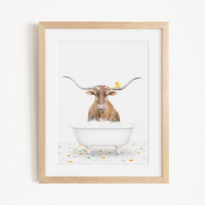 A framed image depicting a brown cow with large horns sitting in a white bathtub, with a yellow duck perched on its head.