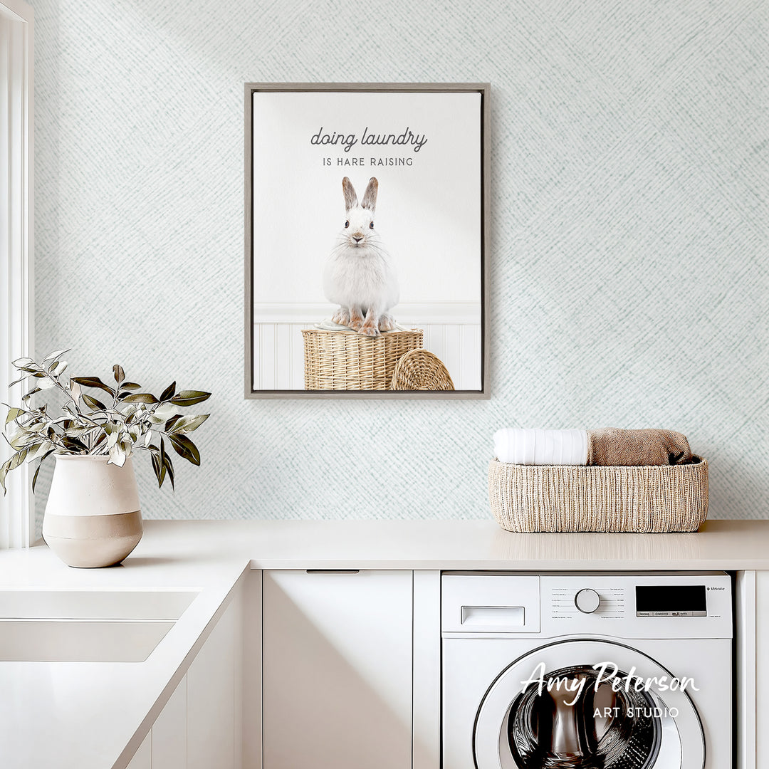 a laundry room with a framed poster of a rabbit sitting on a basket, a potted plant, and a basket of folded towels on a counter.