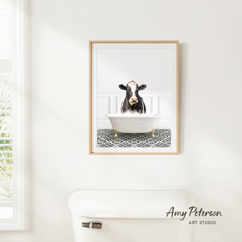A framed image of a cow sitting in a bathtub is hanging on a white wall.