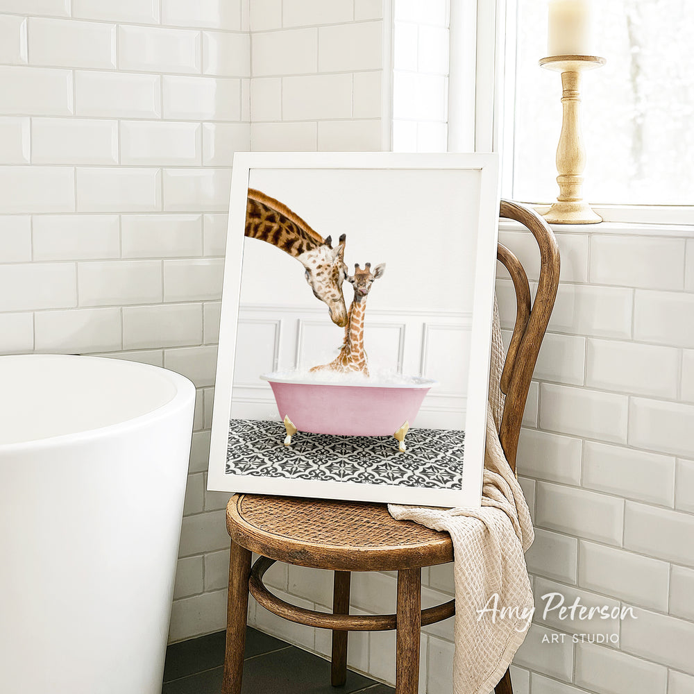A framed image of a giraffe and a baby giraffe sitting in a pink bathtub, with a towel draped over the back of a wooden chair in a bathroom setting.