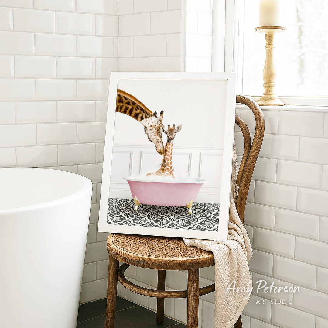 A framed image of a giraffe and a baby giraffe sitting in a pink bathtub, with a towel draped over the back of a wooden chair in a bathroom setting.