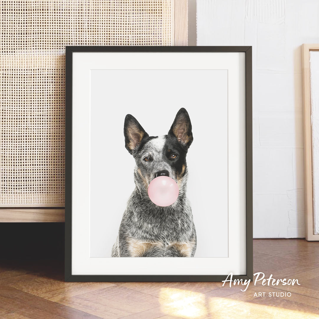 A framed photograph of a dog blowing a bubble with a pink bubble gum bubble.
