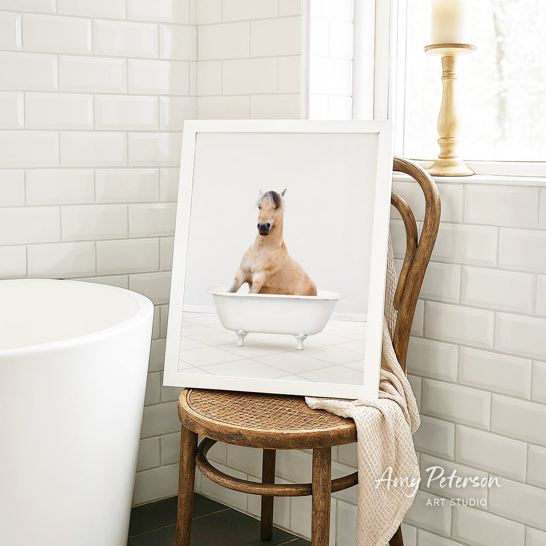 A framed image of a brown horse sitting in a white bathtub, with a towel draped over the back of a wooden chair in a bathroom setting.