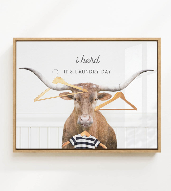 A framed print featuring a bull with a shirt on its back, surrounded by clothes hangers, with the text "i herd, it's laundry day" displayed above the bull.