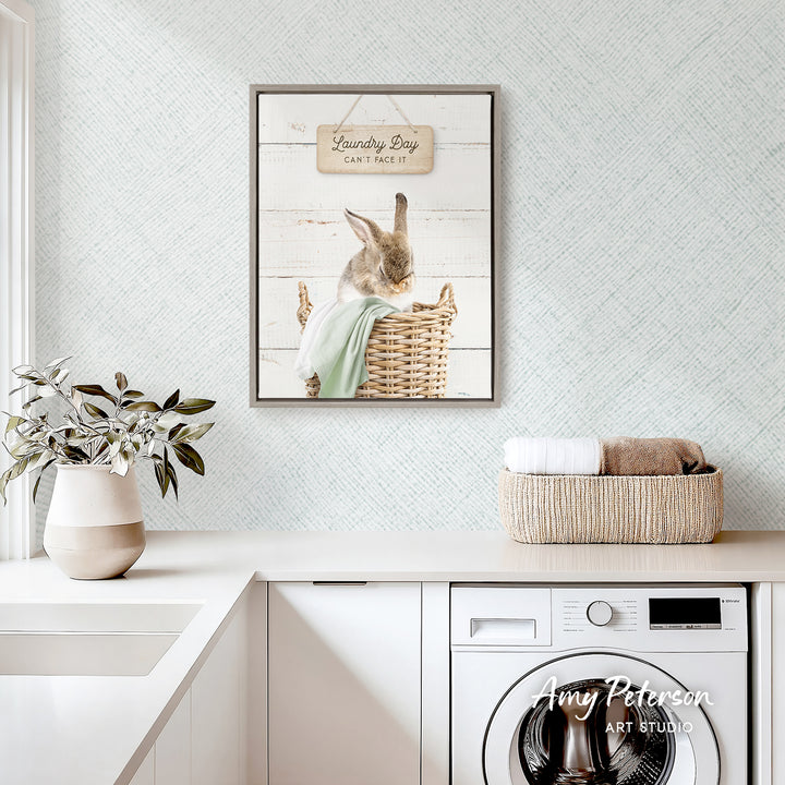 a laundry room with a framed picture of a rabbit sitting in a basket, a potted plant, and a basket of folded towels on a counter.