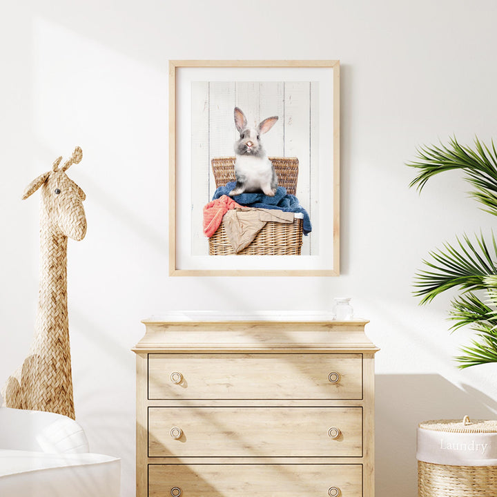 a wooden dresser with a framed picture of a rabbit sitting in a wicker basket on top of it. The dresser is situated in a room with a white wall and a potted plant nearby.
