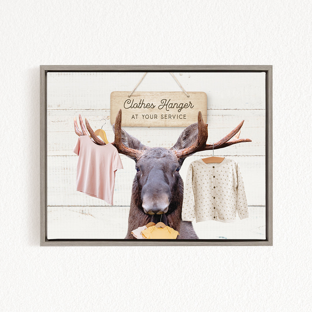 A framed image depicting a moose with antlers holding a clothes hanger sign that reads "Clothes Hanger at Your Service".