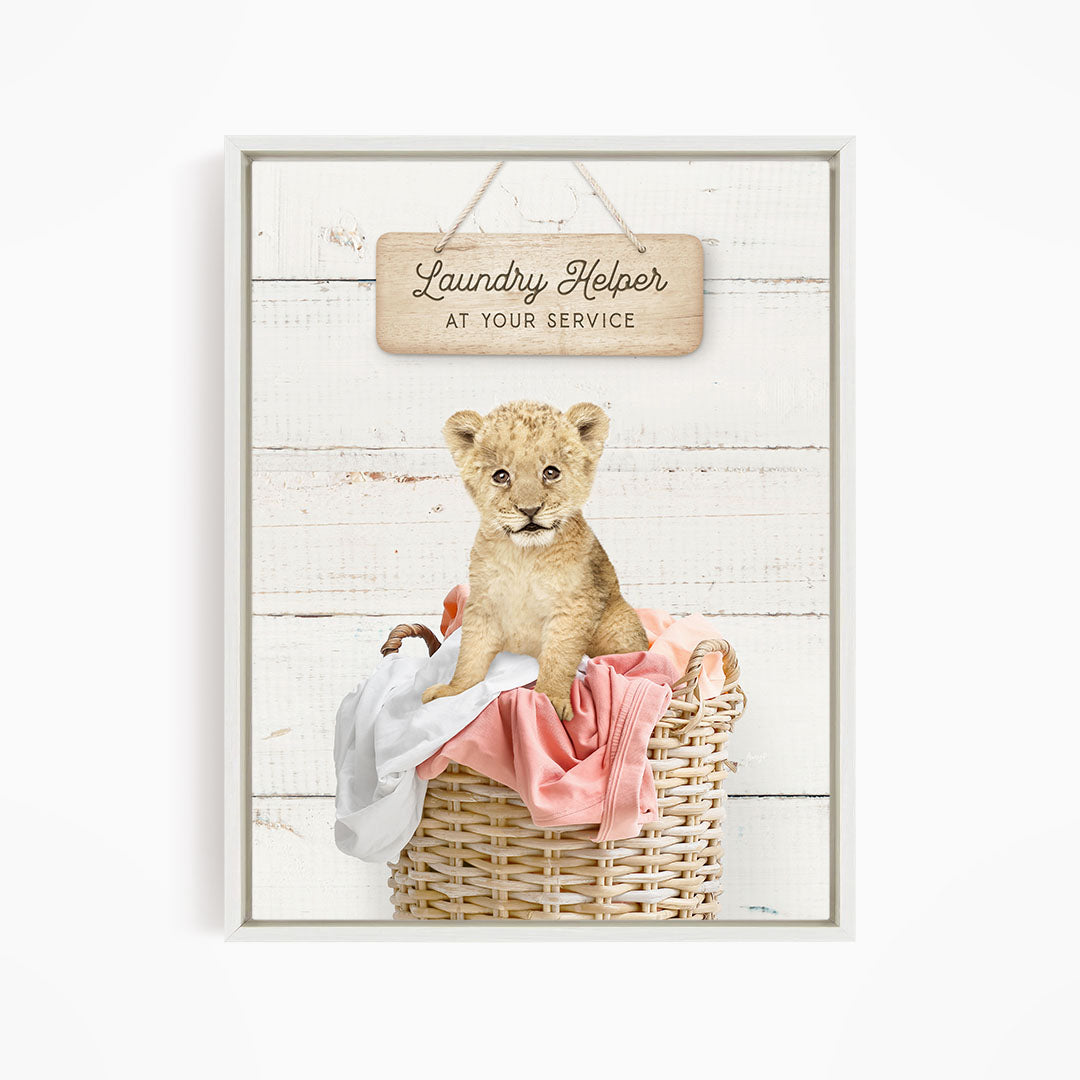 A young lion cub sitting in a wicker basket filled with clothes, with a sign above that reads "Laundry Helper at your service".