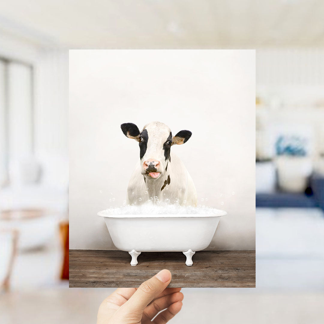 A hand holds up a framed image of a cow sitting in a bathtub filled with water, creating a whimsical and playful scene.