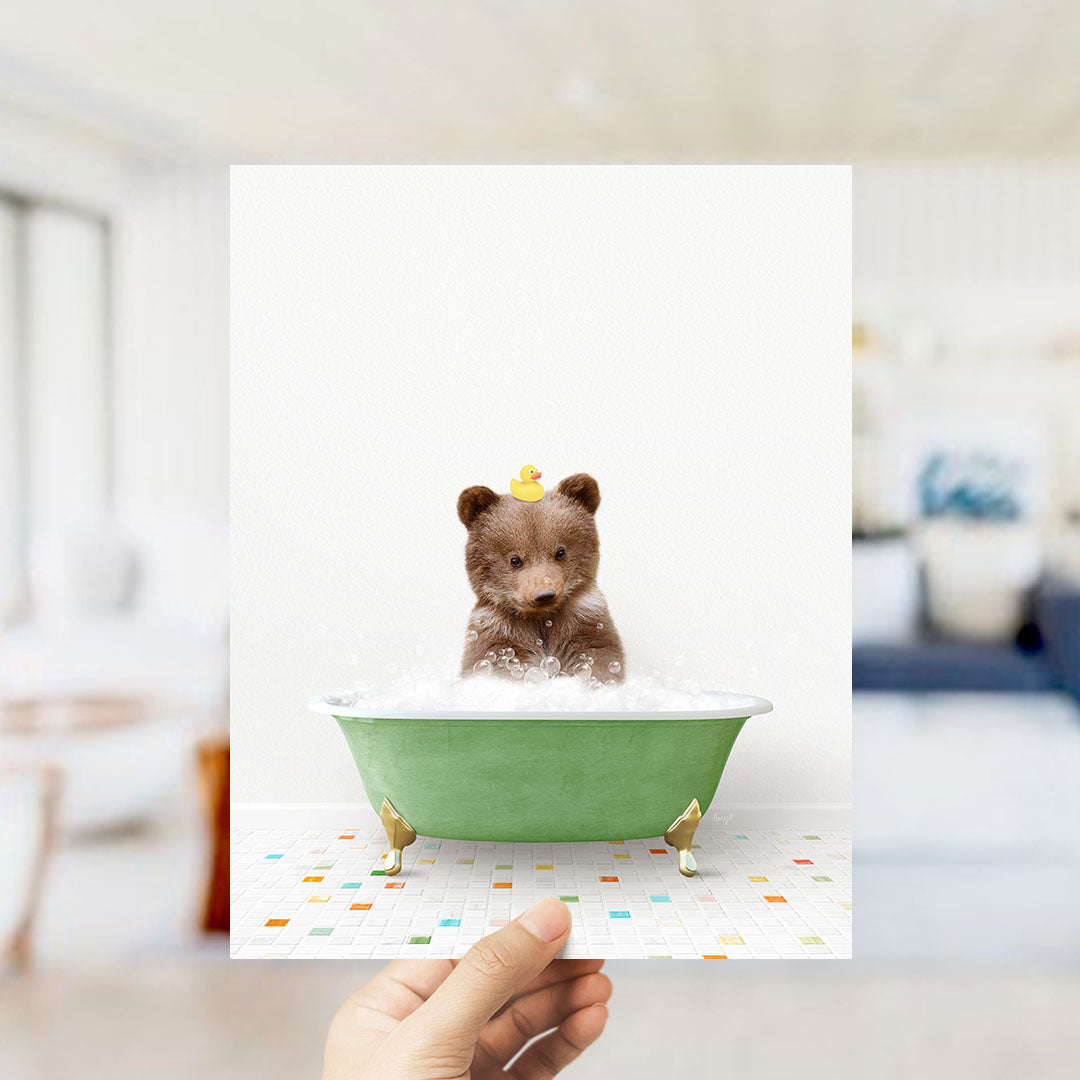 A hand holds up a framed image of a small brown bear sitting in a green bathtub filled with bubbles, with a yellow rubber duck perched on its head.