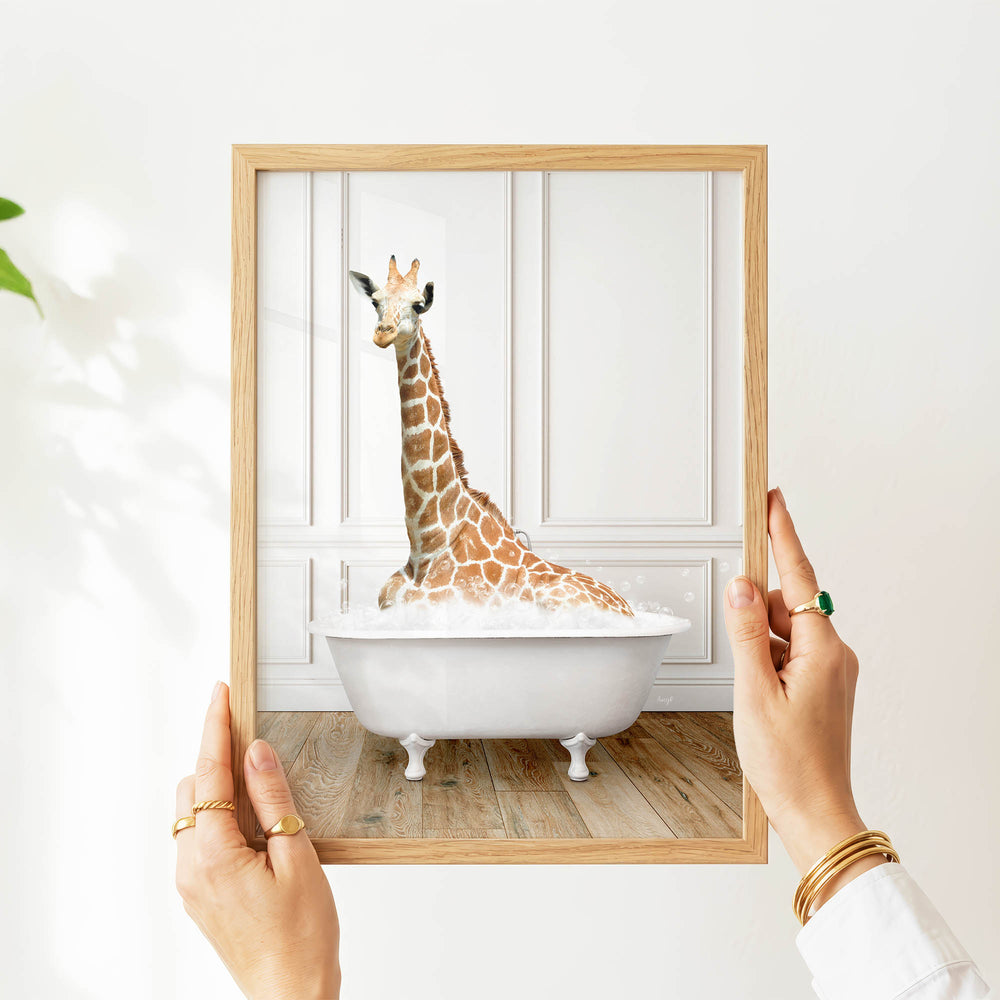 A person's hands are holding a picture frame containing a photograph of a giraffe sitting in a bathtub.
