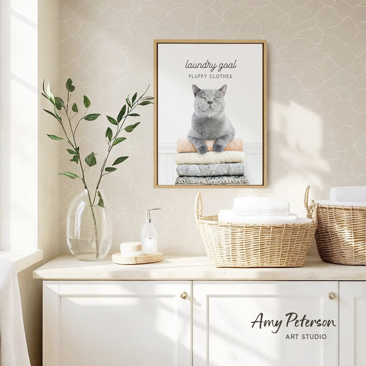 a framed poster of a cat sitting on a pile of laundry, with the text "laundry goal fluffy clothes" and " Amy Peterson ART STUDIO" below it. The poster is displayed on a wall above a white cabinet, which has a wicker basket and a vase with a plant on it.